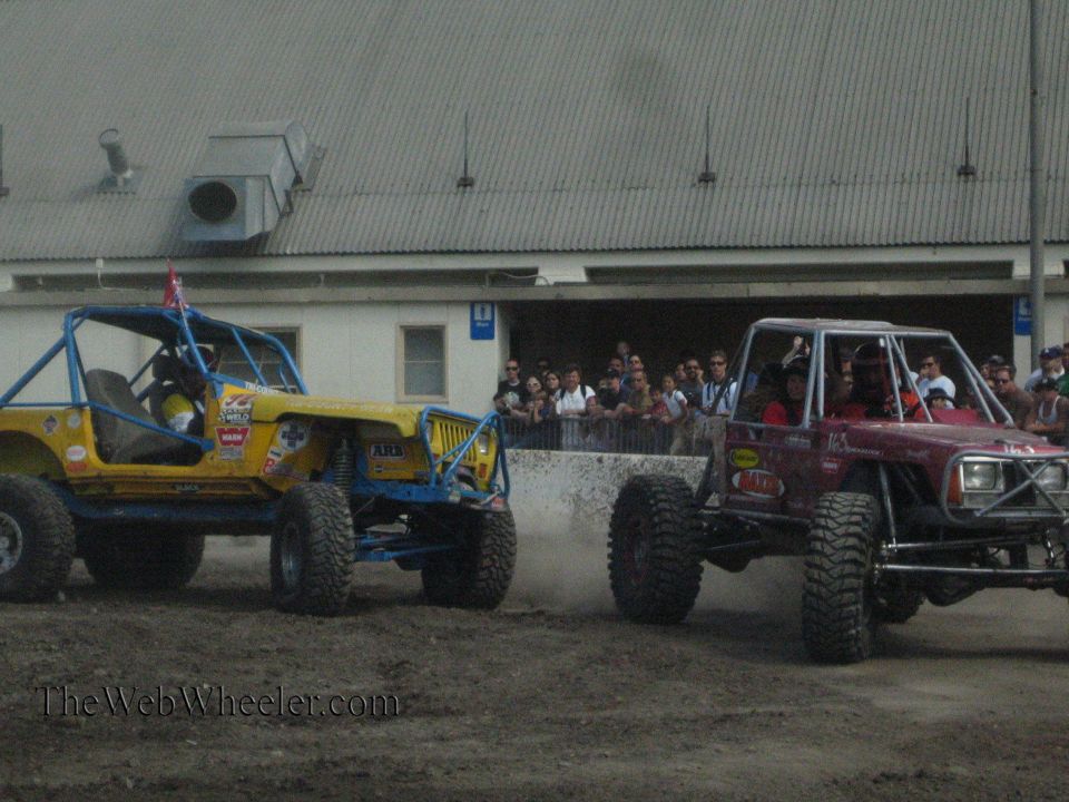 Yellow buggy and maroon buggy on a dirt course with a crowd of spectators behind