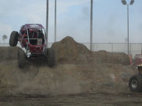 1990 Ford Bronco - maroon tube-chassis rock buggy climbs a large dirt mound, kicking up dust, with a yellow buggy partly visible at right.