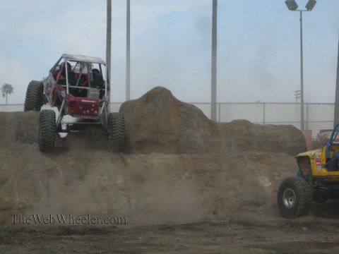 1990 Ford Bronco - maroon tube-chassis rock buggy descends a steep dirt mound while a yellow buggy climbs the slope at right.