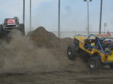 1990 Ford Bronco - maroon rock buggy and a yellow rock buggy raise thick clouds of dust as they work a dirt mound course outdoors.