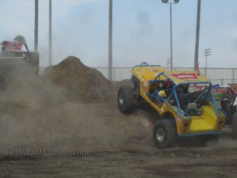 1990 Ford Bronco - yellow tube-chassis rock buggy drives across a dusty dirt course with another buggy and a dirt mound behind it.