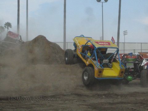 1990 Ford Bronco - yellow tube-chassis buggy with Warn decals drives across a dusty dirt course alongside other rock buggies and a dirt mound.
