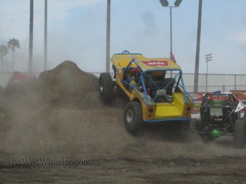 1990 Ford Bronco - yellow tube-chassis rock buggy with Warn decals climbs a dirt mound, kicking up dust, with other buggies in the background.