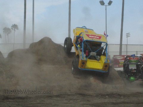1990 Ford Bronco - yellow tube-chassis rock buggy with Warn decals climbs a dirt mound amid heavy dust, with palm trees and stadium lights behind.