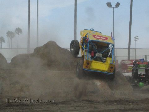 1990 Ford Bronco - yellow tube-chassis rock buggy with Warn decals climbs a dirt mound front-on while another buggy works the course at right.