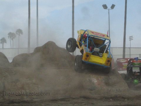 1990 Ford Bronco - yellow tube-frame rock crawler with a spare tire climbs a steep dirt mound during an indoor-style arena competition.