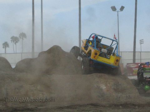 1990 Ford Bronco - yellow rock crawler with a blue roll cage works down a dirt obstacle amid dust at an arena event.