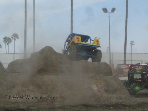 1990 Ford Bronco - blue and yellow tube-frame rock crawler balances on top of a dirt mound in a dusty competition arena.