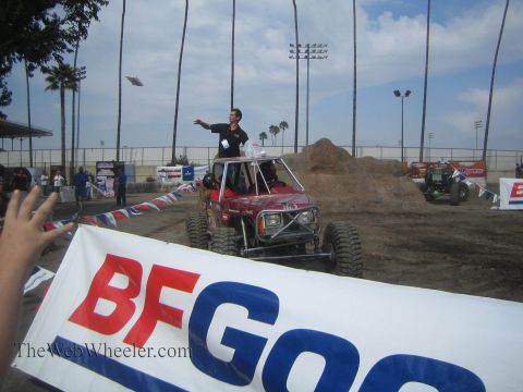 1990 Ford Bronco - man stands up in a red off-road buggy and waves at the crowd behind a large BFGoodrich banner at a dirt arena.