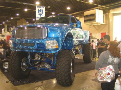 1990 Ford Bronco - heavily lifted blue dually pickup on oversized mud tires sits on display as crowds pass at an indoor expo.