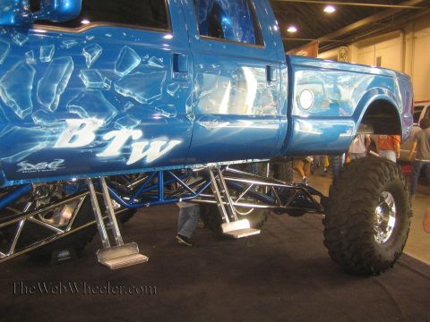 1990 Ford Bronco - side of a lifted blue show truck displays a white BTW graphic above an exposed tubular suspension on chrome steps.