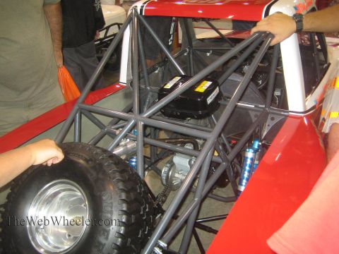 1990 Ford Bronco - People work around the bare tubular chassis of a red off-road race truck showing the battery, shocks and a spare tire.
