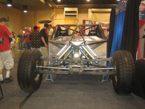 1990 Ford Bronco - Front view of a custom tube-chassis buggy on stands, displaying its exposed V engine and twin coilover shocks at an indoor expo.