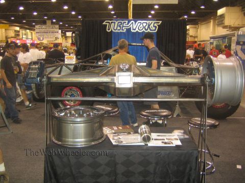 1990 Ford Bronco - bare tubular chassis hangs on display at a Tube-Works booth above off-road wheels and brochures while two men talk behind it.