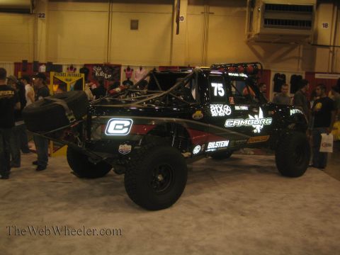 1990 Ford Bronco - black No. 75 Camburg-sponsored off-road race truck with lit number panels sits on display in an indoor expo hall.