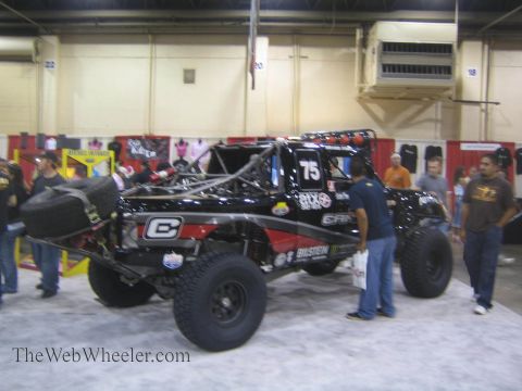 1990 Ford Bronco - Spectators walk around a black No. 75 Camburg off-road race truck displayed on the carpet of an indoor expo hall.