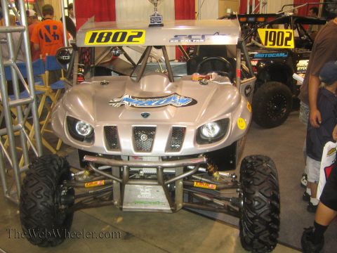 1990 Ford Bronco - silver Yamaha Rhino-style race side-by-side marked No. 1802 sits on display with another UTV behind it at an indoor expo.