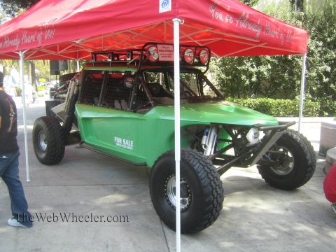 1990 Ford Bronco - green open tube-frame off-road buggy with a For Sale sign sits under a red pop-up canopy on a paved outdoor display.