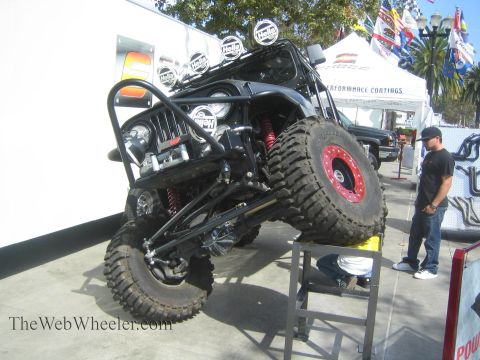 1990 Ford Bronco - black tube-frame rock buggy is posed with its front wheel lifted, exposing a front winch and red coilover shocks at an outdoor booth.