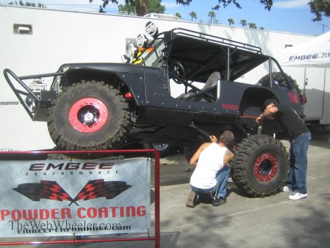 1990 Ford Bronco - matte-black open-body Jeep-style rock crawler with a roll cage sits raised on red beadlock wheels while two men work on it beside an Embee…