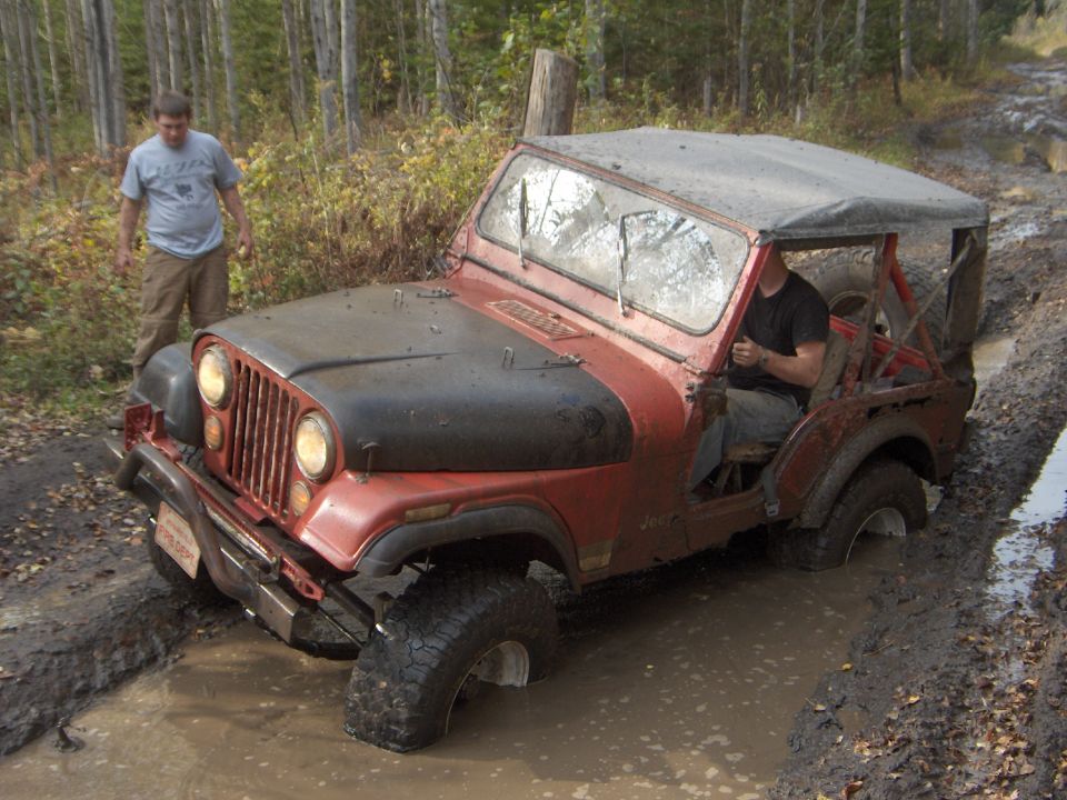 Muddy red Jeep CJ with winch bumper and soft top stuck in a rutted mud hole on a forest trail
