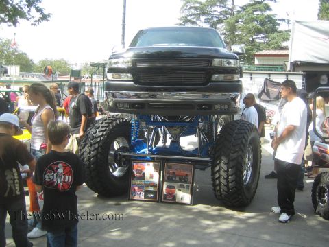 1990 Ford Bronco - black Chevrolet pickup sits sky-high on a blue tubular suspension and massive mud tires, with framed award photos propped beneath it at an…