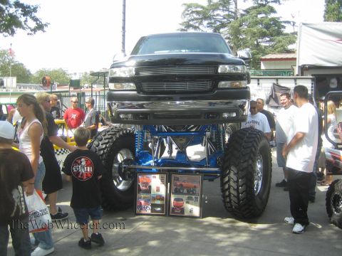 1990 Ford Bronco - Crowd gathers around a lifted black Chevrolet pickup raised on a bright blue tube suspension over huge mud tires with framed photos display…