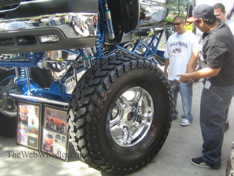 1990 Ford Bronco - polished chrome wheel wrapped in a mud-terrain tire sits under a blue tubular suspension as two men inspect the lifted truck at a show.