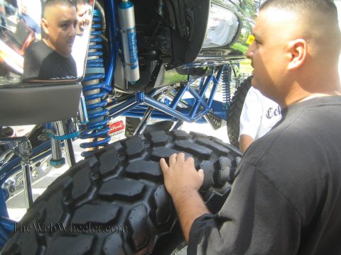 1990 Ford Bronco - man in black grips a mud-terrain tire while inspecting the blue coilover suspension and chrome links of a lifted show truck.