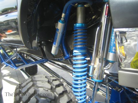 1990 Ford Bronco - Detail of bright blue coilover shocks, springs and tubular control arms with a polished exhaust under the body of a lifted show truck.