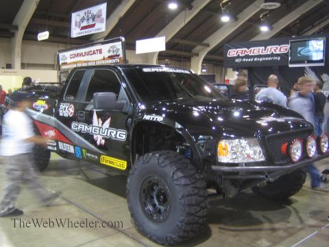 1990 Ford Bronco - black off-road prerunner truck wearing Camburg, Nitto and Bilstein graphics with roof and bumper lights is displayed at an indoor trade sho…