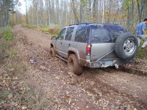2000 Chevrolet Tahoe - An SUV is stuck in deep mud on a wooded dirt road, with a person walking in the background.