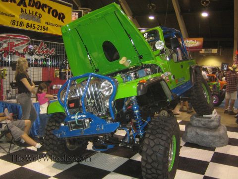 1990 Ford Bronco - bright green tube-bodied Jeep rock crawler with its hood open and blue front suspension sits on a checkered display floor at an indoor show.