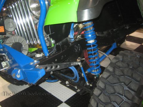 1990 Ford Bronco - Detail of a green off-road vehicle's front end showing blue coilover shock, tubular links, leaf spring and a mud tire over a checkered floo…