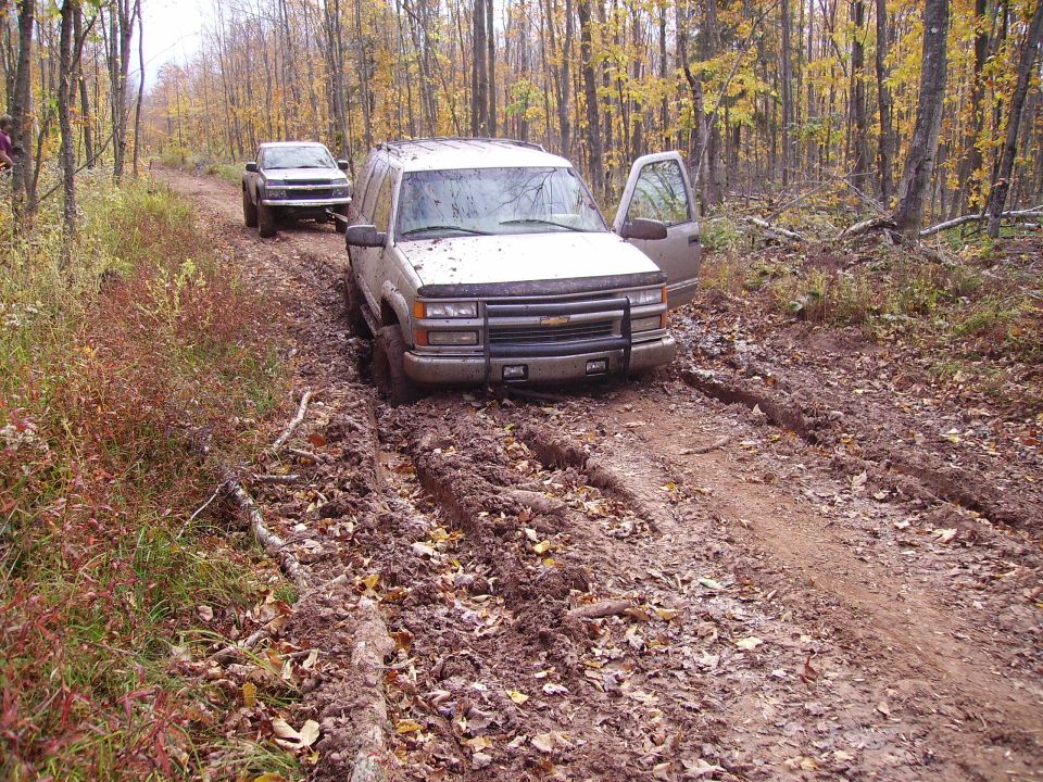 Silver Chevrolet Tahoe stuck in mud on a forest road, driver's door open