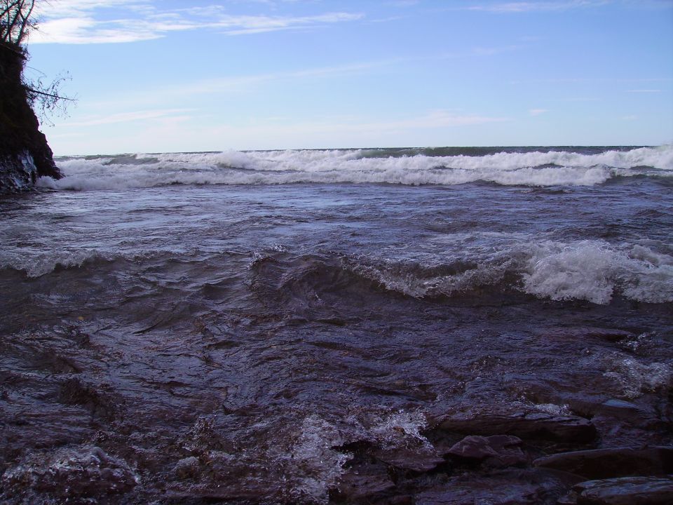 Ocean waves crash on a rocky shore under a blue sky with scattered clouds. A watermark reads SUPERMOTORS