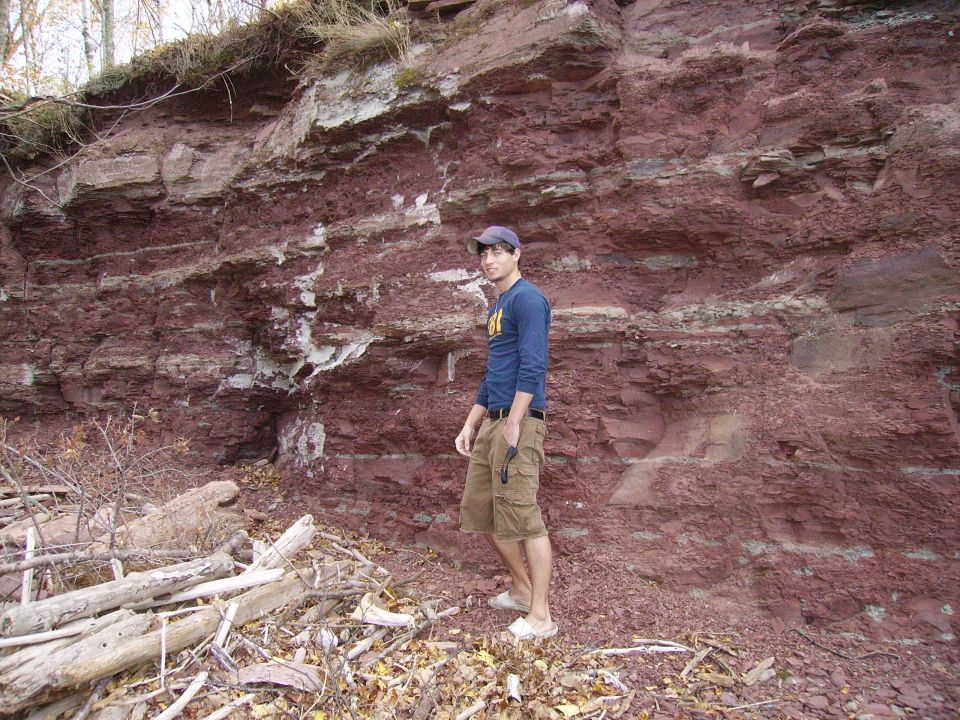 Man in blue shirt and cargo shorts stands by a layered red rock cliff face with fallen branches in the foreground. SuperMotors.net logo visible