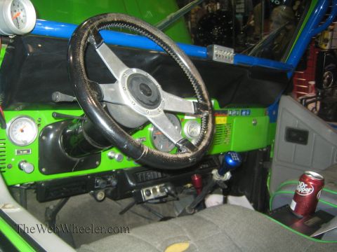 1990 Ford Bronco - Interior of a green caged Jeep showing a worn black steering wheel, round gauges, floor controls and a Dr Pepper can in the door pocket.