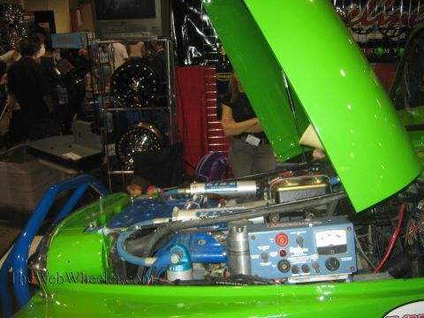 1990 Ford Bronco - bright green hood is propped open over an engine bay fitted with hydraulic-ram steering and a control box at an indoor display booth.