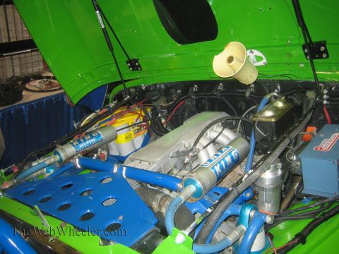 1990 Ford Bronco - Engine bay of a green off-road vehicle showing a V-engine, King reservoir shocks, yellow battery and a blue lightening-holed mounting plate.
