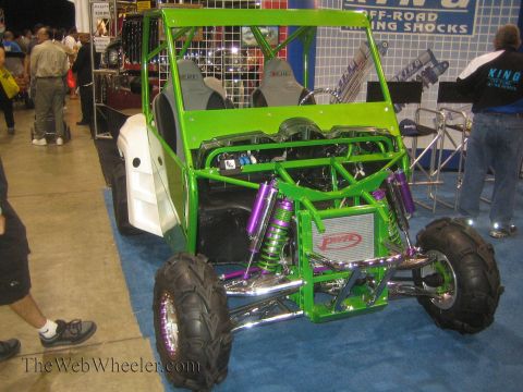 1990 Ford Bronco - green tube-chassis side-by-side buggy with purple coilover shocks and chrome suspension is displayed on blue carpet at a King Shocks booth.