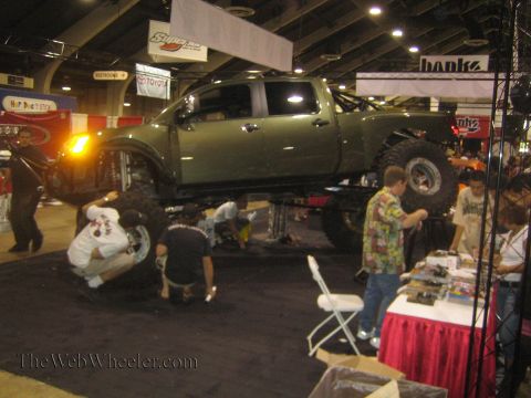 1990 Ford Bronco - dark green crew-cab prerunner pickup sits raised on jack stands while crew work underneath it at a busy indoor trade-show booth.