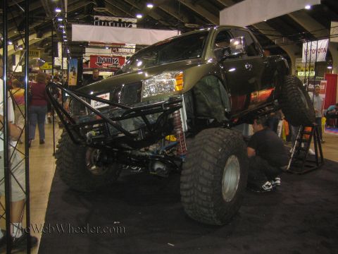 1990 Ford Bronco - black Nissan crew-cab pickup built as a crawler with a tubular front bumper and big tires sits on a jack stand as a man works beneath it.