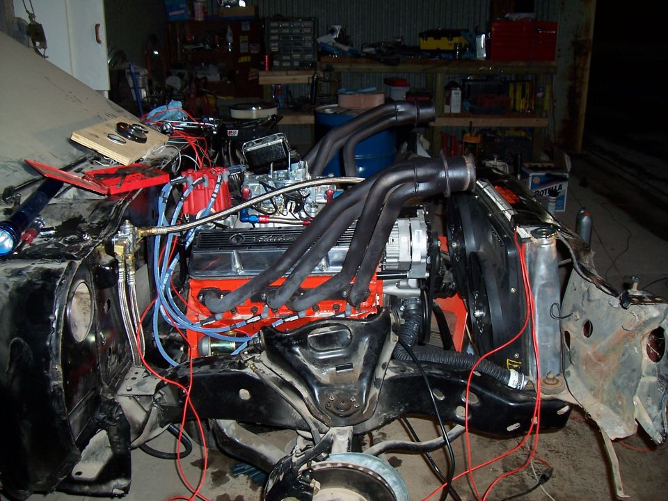 Engine bay of a car with a V8 engine, headers, and wiring. The front end of the car is disassembled