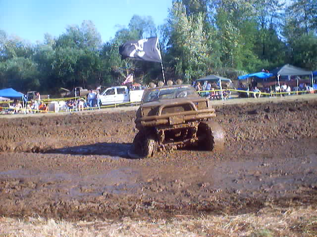 Mud bogging truck with a skull flag driving through mud; spectators and tents in background