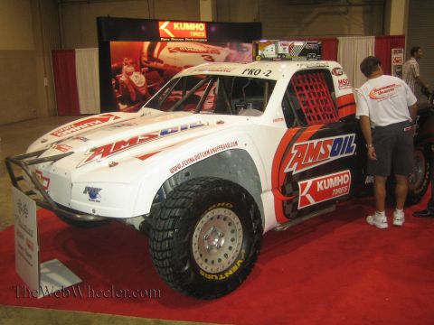 1990 Ford Bronco - white off-road race truck wrapped in AMSOIL, Kumho Tires and PRO-2 graphics sits on a red carpet display at an indoor expo.