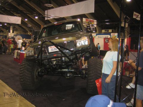 1990 Ford Bronco - dark lifted pickup on oversized mud tires with a tube front bumper sits on display while a woman looks on at an indoor expo.