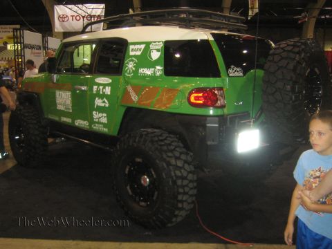 1990 Ford Bronco - Rear three-quarter view of a green-and-white lifted Toyota FJ Cruiser on mud tires with a rear-mounted spare at an indoor show.