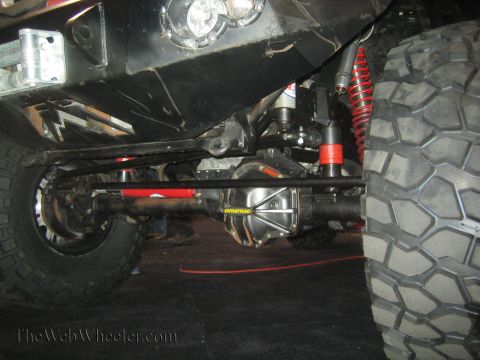 1990 Ford Bronco - Close-up underbody view of a vehicle's front solid axle, driveshaft and red coilover shocks beside a knobby off-road tire.
