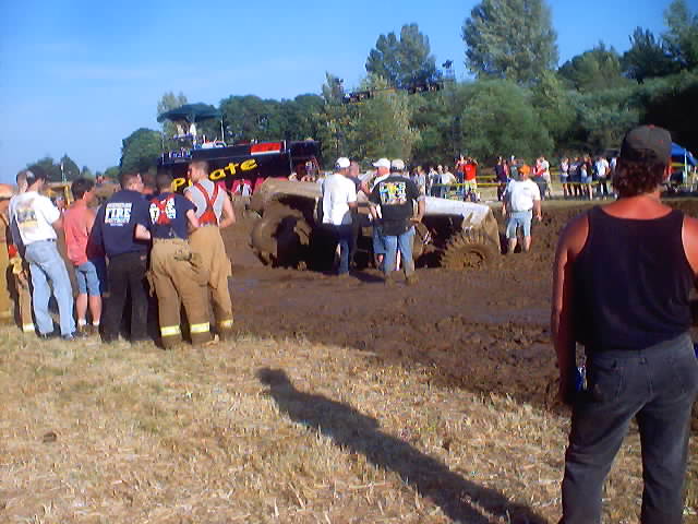 People watching a mud bogging event with a truck stuck in the mud. A large black and red vehicle is visible in the background